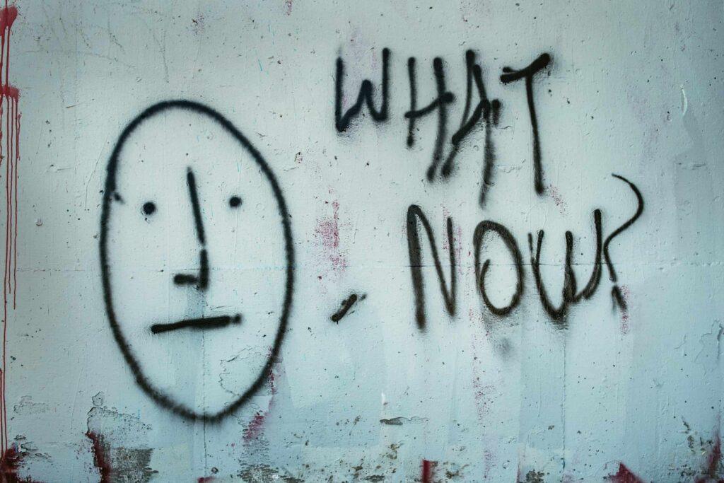 Graffiti in black in on a pale wall Says "what now" with a simple drawing of a face looking expressinless