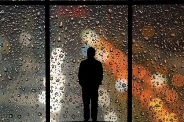 The silhouette of a man looking through a large window, into the city below representing the isolation and loneliness he feels due to his chronic illness